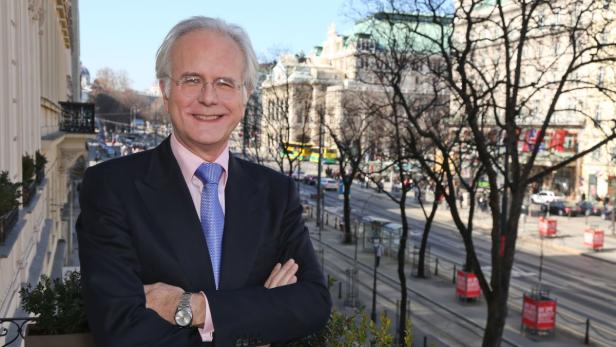Ein lächelnder Mann im Anzug steht vor einer Wiener Stadtlandschaft.
