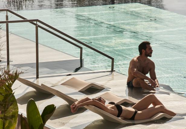 Eine Frau entspannt auf einer Liege am Pool, während ein Mann daneben sitzt.
