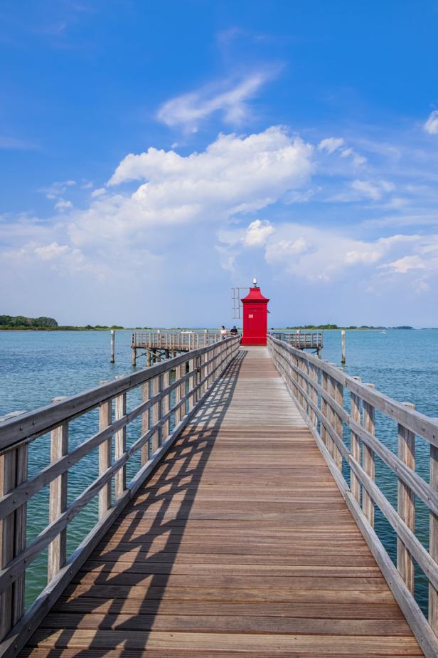 Ein Holzsteg führt zu einem roten Leuchtturm am Meer unter blauem Himmel.