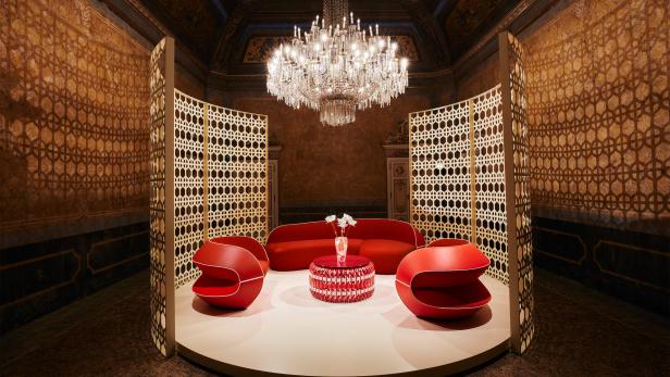 Ein rotes Sofa und Sessel stehen unter einem Kronleuchter in einem opulenten Raum.