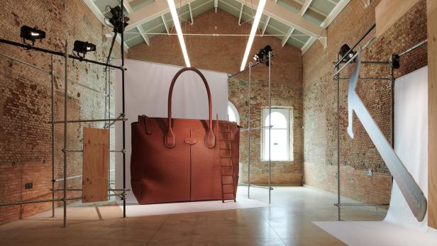 Eine übergroße braune Tod's Handtasche steht in einem Raum mit Backsteinwänden und Baugerüsten.