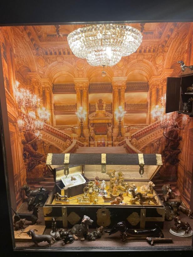 Eine Sammlung von Dackel-Figuren in einer offenen Truhe vor einer opulenten Palastkulisse.