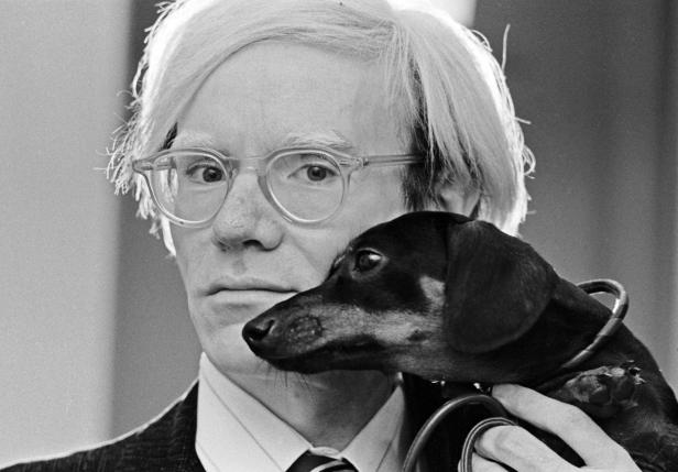 Andy Warhol mit Brille hält einen Dackel im Arm.