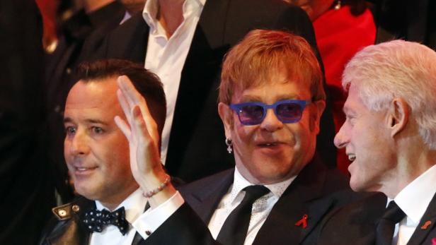 David Furnish, Elton John mit blauer Brille und Bill Clinton sitzen nebeneinander.