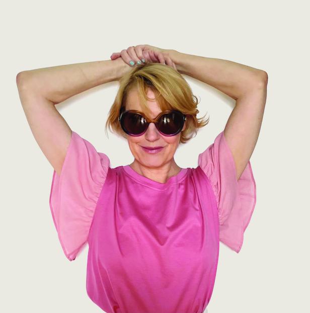 Eine Frau mit blonden Haaren und runder Sonnenbrille posiert in einem rosa Oberteil.