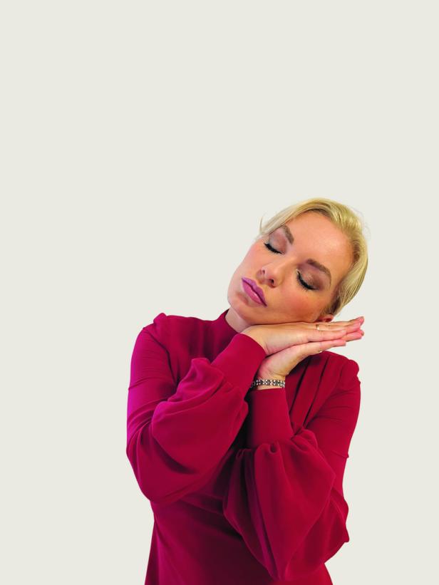 Eine blonde Frau in einem roten Kleid ruht mit geschlossenen Augen und verschränkten Händen.