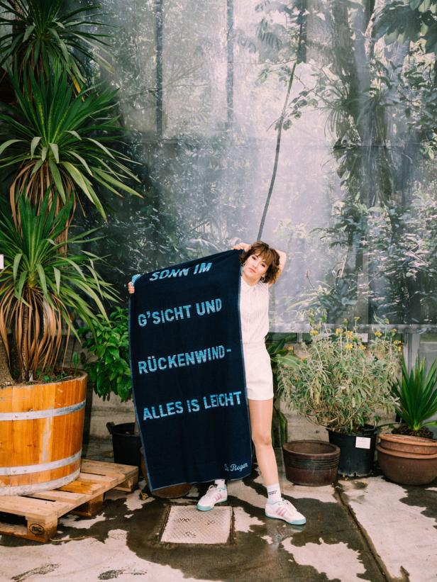 Eine Frau hält ein blaues Handtuch mit der Aufschrift „Sonn im G’sicht und Rückenwind – Alles ist leicht“ in einem Gewächshaus.