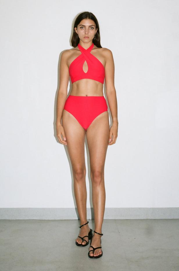 Eine Frau steht in einem roten Bikini und schwarzen Sandalen vor einer weißen Wand.