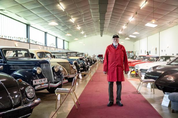 Ein Mann in rotem Mantel steht in einer Halle voller Oldtimer.