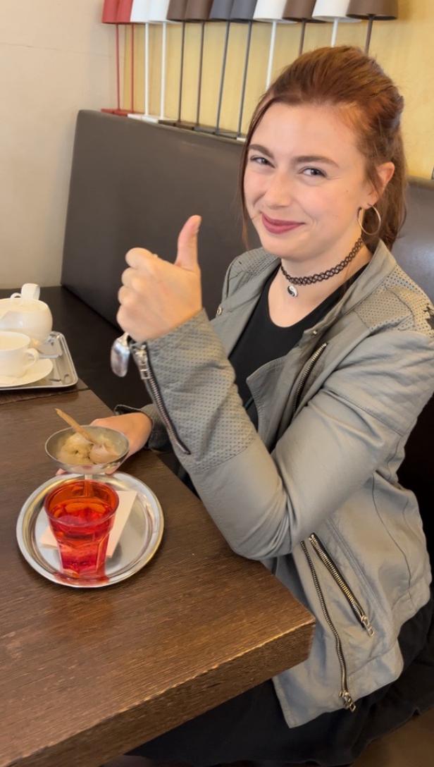 Eine Frau sitzt in einem Café und gibt einen Daumen nach oben, während sie ein Getränk und Eis genießt.