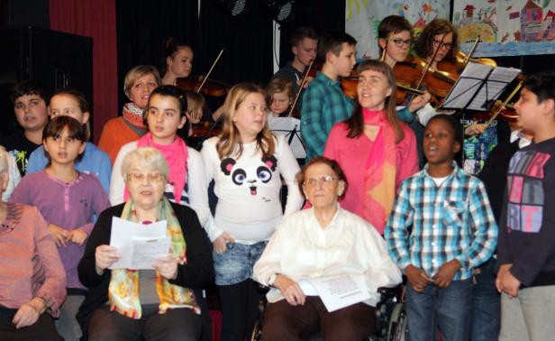 Eine Gruppe von Kindern und älteren Menschen singt und spielt Geige auf einer Bühne.
