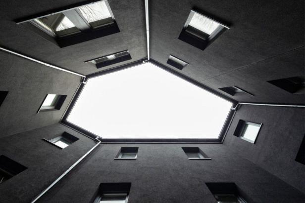 Blick von unten in einen Innenhof mit dunklen Wänden und mehreren Fenstern zum Himmel.