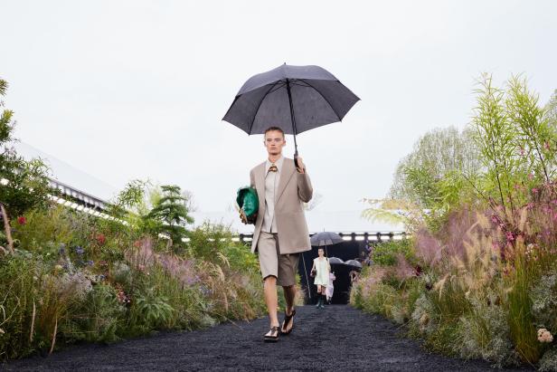 Ein Model mit Schirm und grüner Tasche auf einem Laufsteg mit Blumen und Pflanzen.