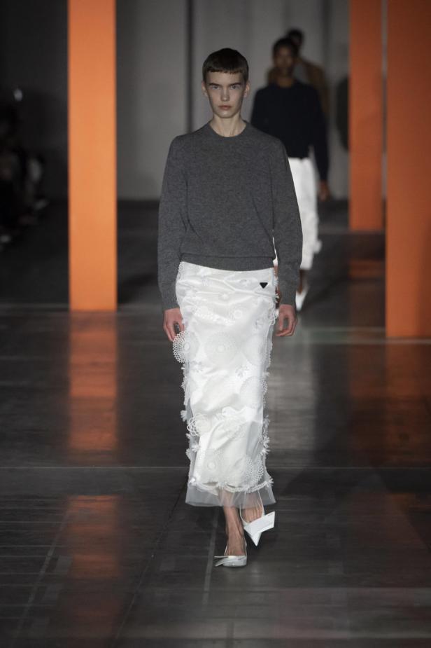 Ein Model auf dem Laufsteg, gekleidet in einen grauen Pullover und einen langen, weißen Rock mit Spitzenapplikationen.