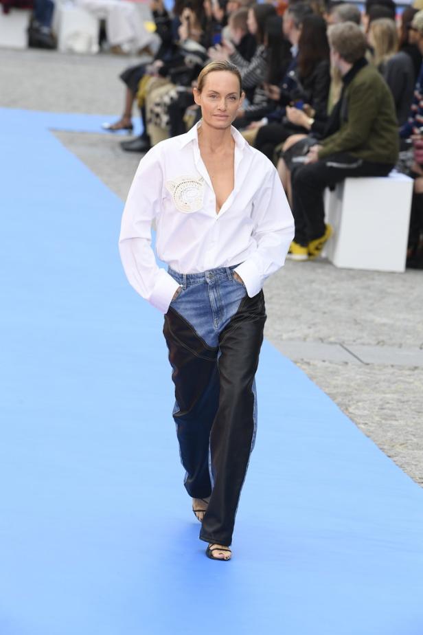 Ein Model läuft in einem weißen Hemd und einer Hose mit Denim- und Ledereinsätzen über einen blauen Laufsteg.