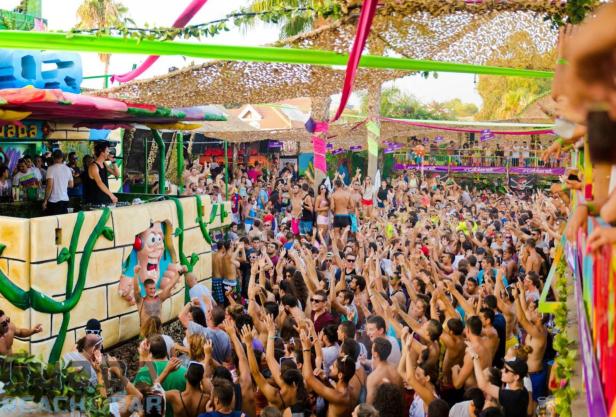 Eine große Menschenmenge feiert auf einer Open-Air-Party mit einem DJ auf einer dekorierten Bühne.