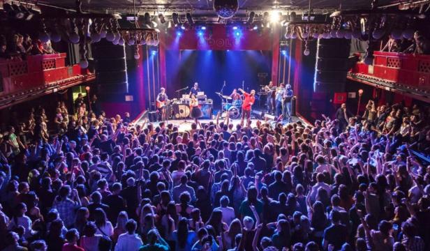 Eine Band spielt vor einem großen, jubelnden Publikum in einem Konzertsaal.