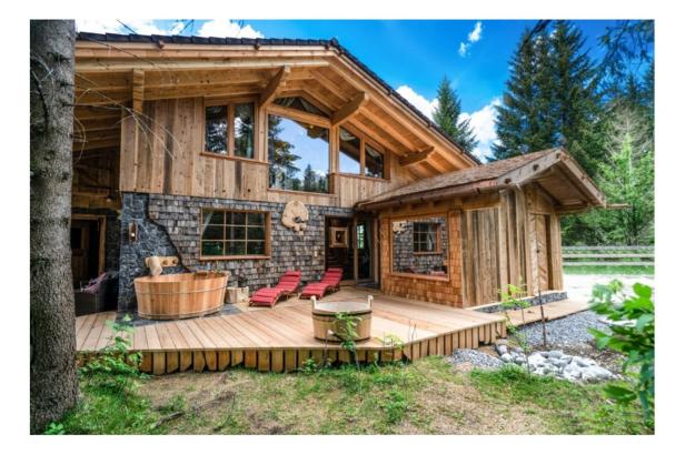Ein Holzhaus mit Terrasse, Whirlpool und Liegestühlen im Grünen.