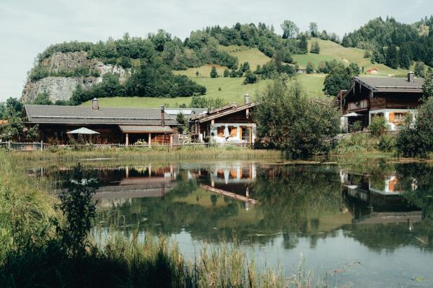 Mehrere Holzhäuser spiegeln sich in einem ruhigen See vor einer grünen Hügellandschaft.