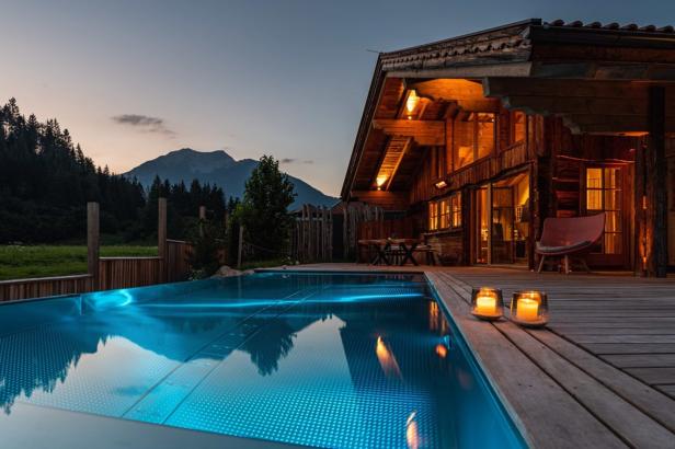 Ein beleuchteter Pool vor einem Holzhaus in den Bergen bei Dämmerung.