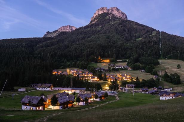 Ein Bergdorf liegt eingebettet in einer grünen Landschaft unter einem hohen Bergmassiv.