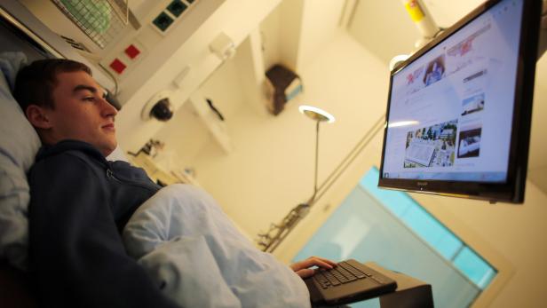 Ein Mann liegt im Bett und bedient einen Laptop, während er auf einen an der Wand montierten Bildschirm schaut.