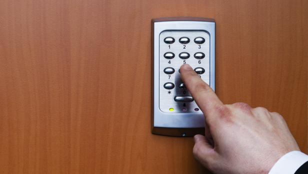 Eine Hand tippt eine „5“ auf einem Zahlenschloss an einer Holztür ein.