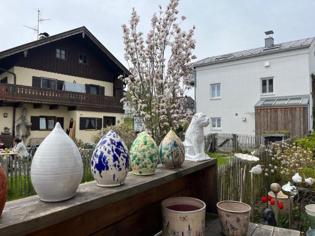 Dekorative Keramikobjekte und Blumentöpfe stehen vor einem Haus mit blühendem Baum.
