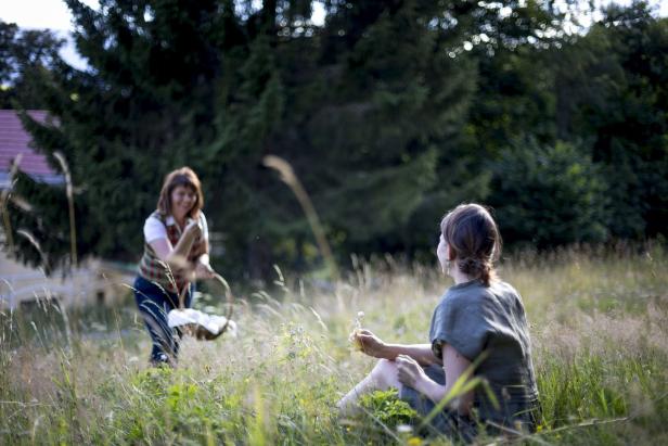 Eine Frau sitzt im Gras, während eine andere mit einem Korb auf sie zukommt.