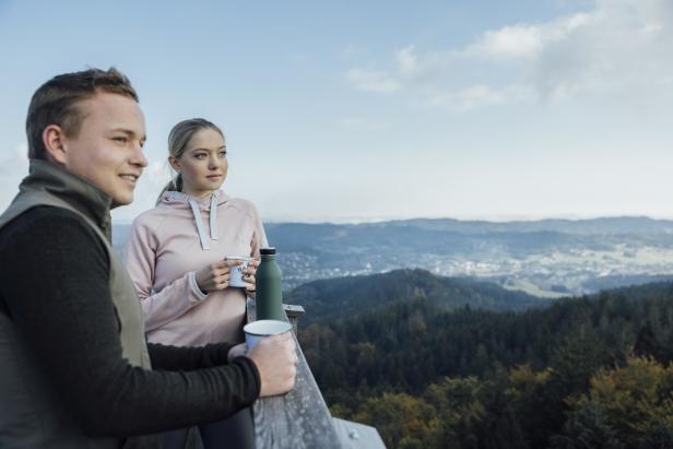 Ein junges Paar genießt die Aussicht von einem Aussichtsturm mit Tassen in der Hand.