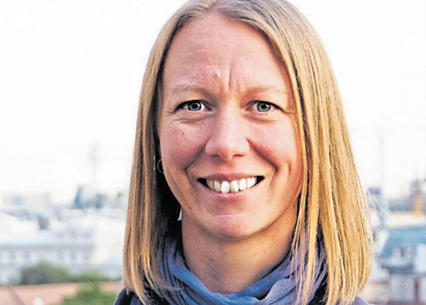 Eine lächelnde Frau mit blonden Haaren und blauem Schal vor einem unscharfen Hintergrund.