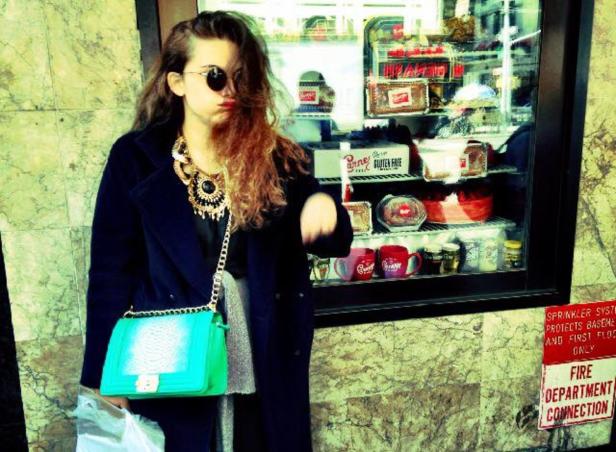 Eine junge Frau mit Sonnenbrille, auffälliger Halskette und grüner Handtasche steht vor einem Schaufenster.