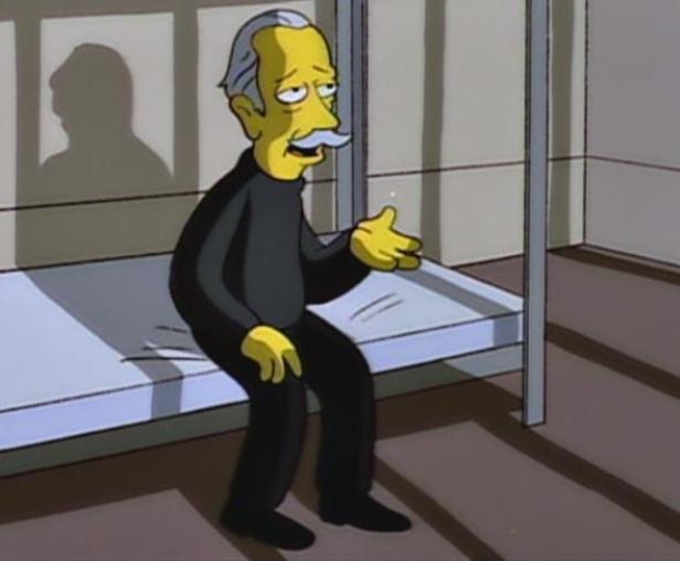 Ein älterer Mann mit Schnurrbart sitzt traurig auf einem Bett in einem kahlen Raum.