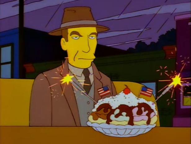 Ein Mann mit Hut und Anzug sitzt vor einem großen Eisbecher mit Wunderkerzen und kleinen US-Flaggen.