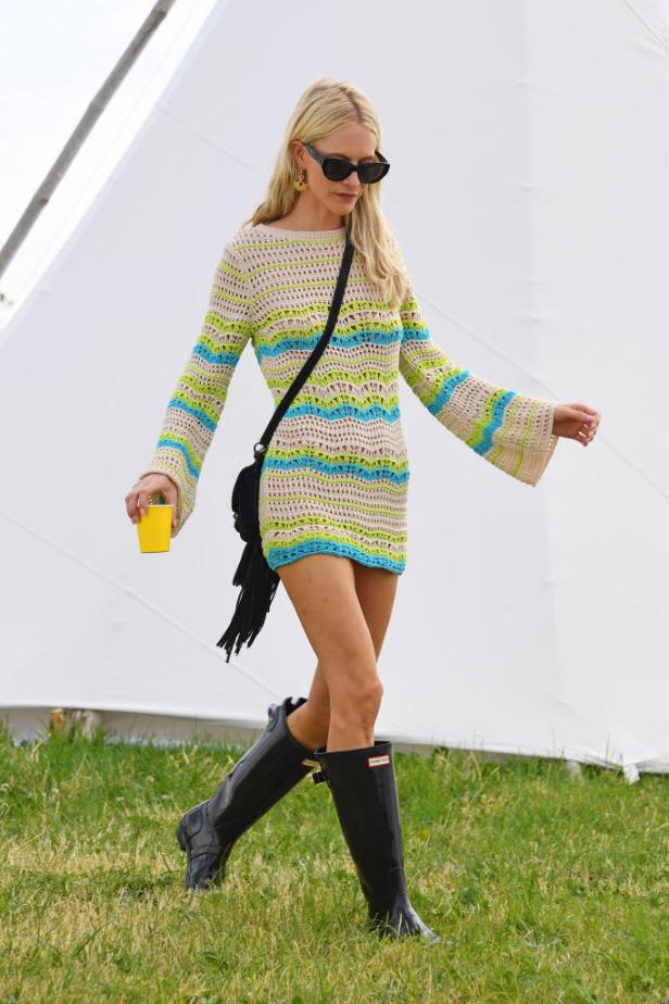 Eine blonde Frau mit Sonnenbrille, Häkelkleid und Gummistiefeln geht über eine Wiese.