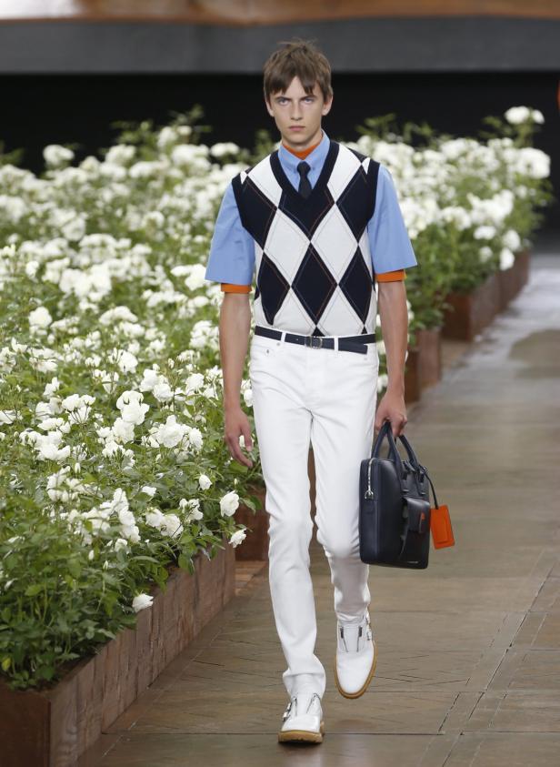 Ein Mann mit Argyle-Weste, Hemd und Krawatte geht mit einer Aktentasche an weißen Rosen vorbei.