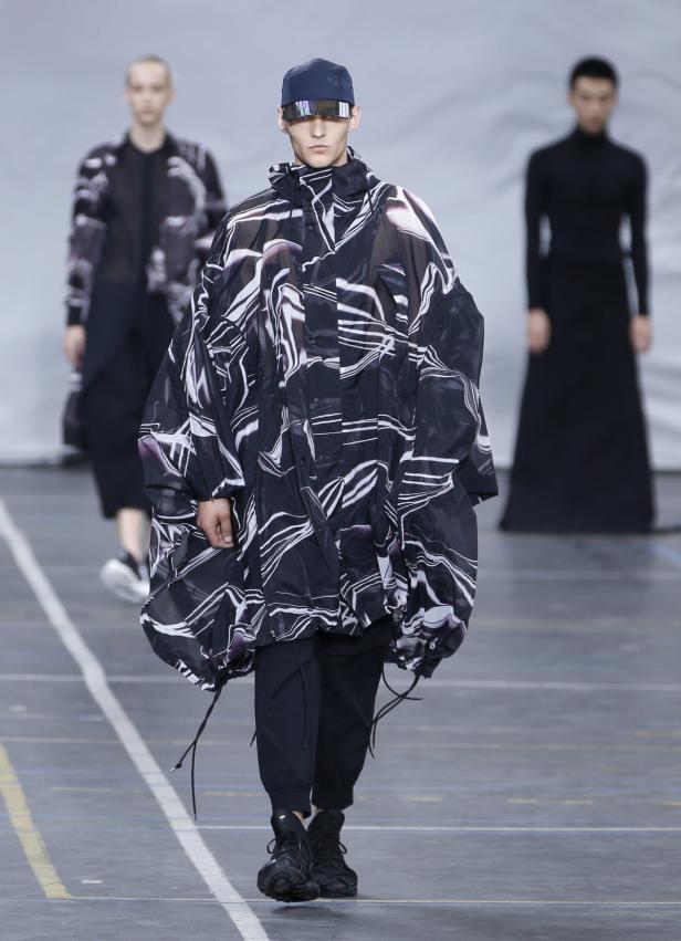 Ein Model auf dem Laufsteg trägt eine übergroße Jacke mit abstraktem Muster und eine futuristische Brille.