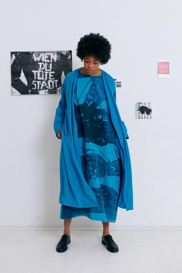 Eine Frau mit Afro trägt ein blaues Kleid und einen passenden Cardigan vor einer weißen Wand mit Bildern.