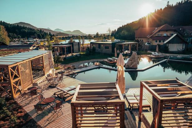 Blick auf ein Resort mit Schwimmteich, Holzhütten und Bergen im Hintergrund.