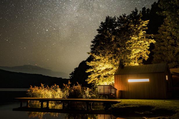 Ein Steg führt zu einem beleuchteten Haus unter einem Sternenhimmel.