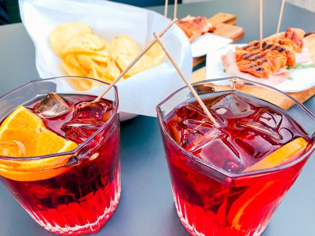 Zwei Gläser Negroni mit Orangenscheiben und Eiswürfeln, dazu Chips und gegrillte Speisen.