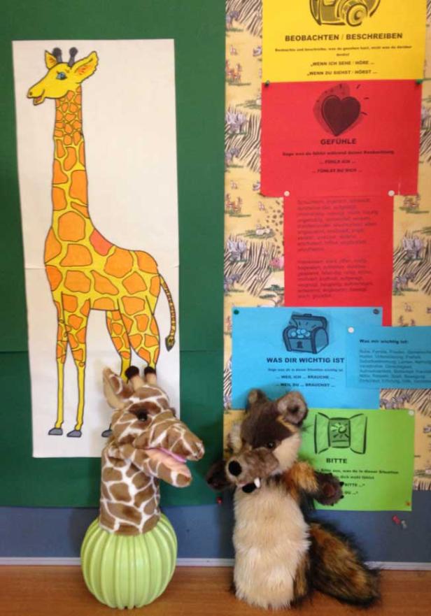 Eine Giraffenzeichnung, eine Giraffenhandpuppe und ein Waschbär-Stofftier vor einer Pinnwand.