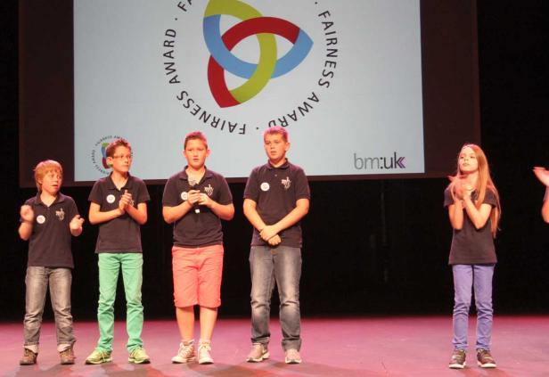 Eine Gruppe Kinder steht auf einer Bühne vor einem Bildschirm mit der Aufschrift „Fairness Award“.