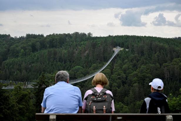 Drei Personen sitzen auf einer Bank und blicken auf eine lange Hängebrücke über einen bewaldeten Hügel.