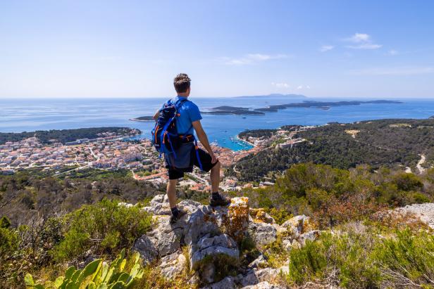 Ein Wanderer blickt von einem Felsen auf eine Küstenstadt und das Meer.