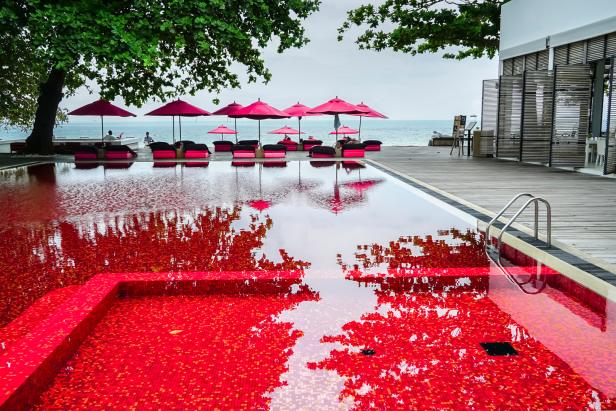 Ein roter Pool spiegelt Bäume und pinke Sonnenschirme an einem Strand wider.