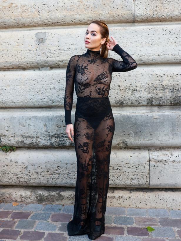 Rita Ora posiert in einem schwarzen, transparenten Spitzenkleid vor einer Steinmauer.
