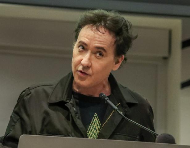 John Cusack spricht bei einer Veranstaltung am Rednerpult.