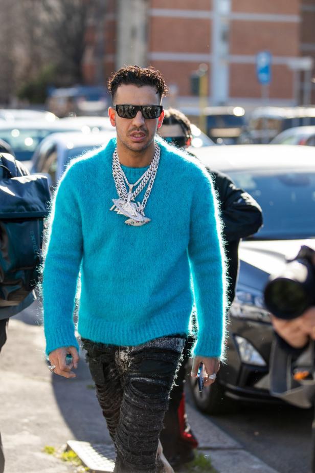 Ein Mann mit Sonnenbrille und auffälligem Schmuck trägt einen türkisfarbenen Pullover.
