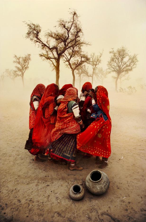 Eine Gruppe indischer Frauen in roten Gewändern steht in einer staubigen Landschaft.